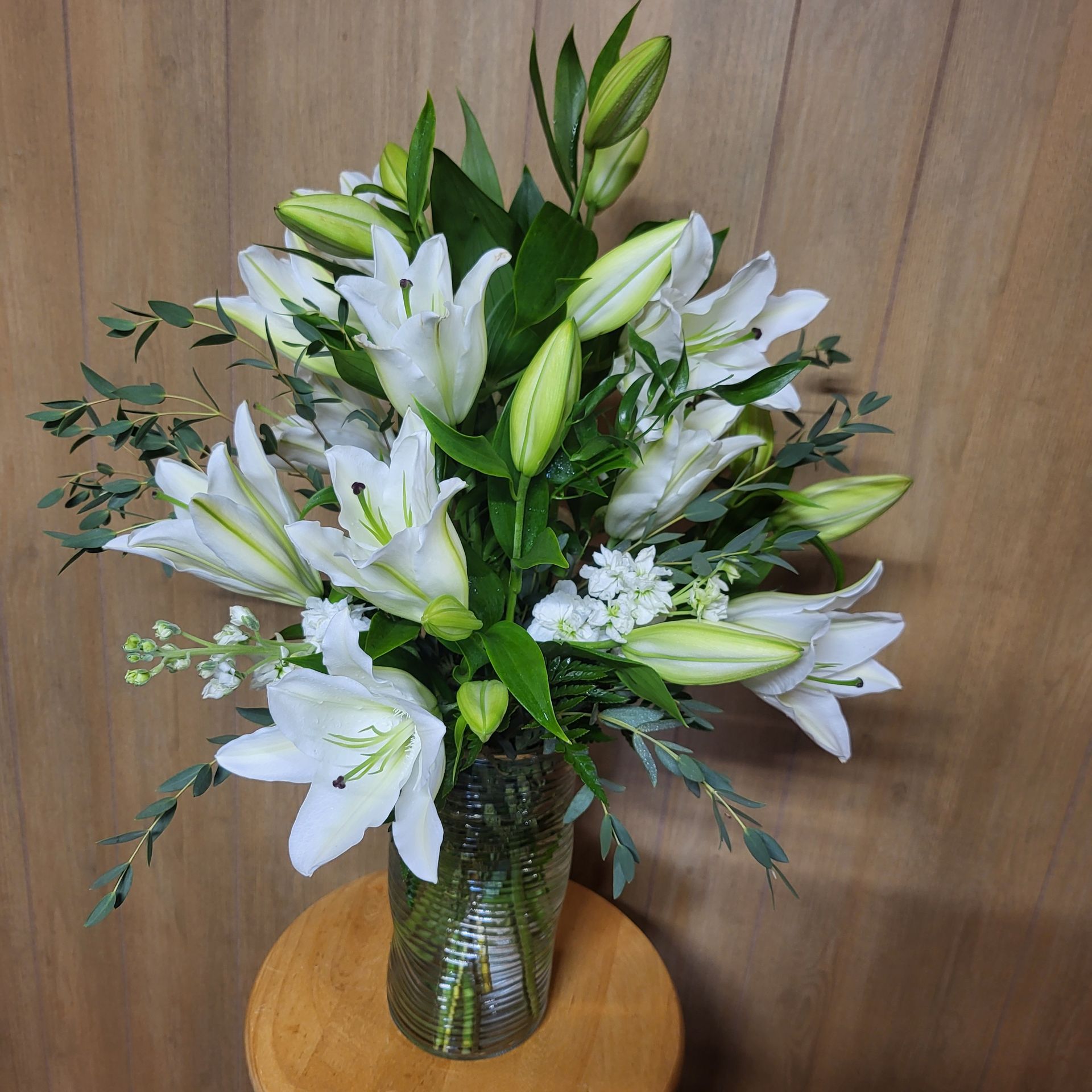 A vase filled with white lilies and greenery is sitting on a wooden table.