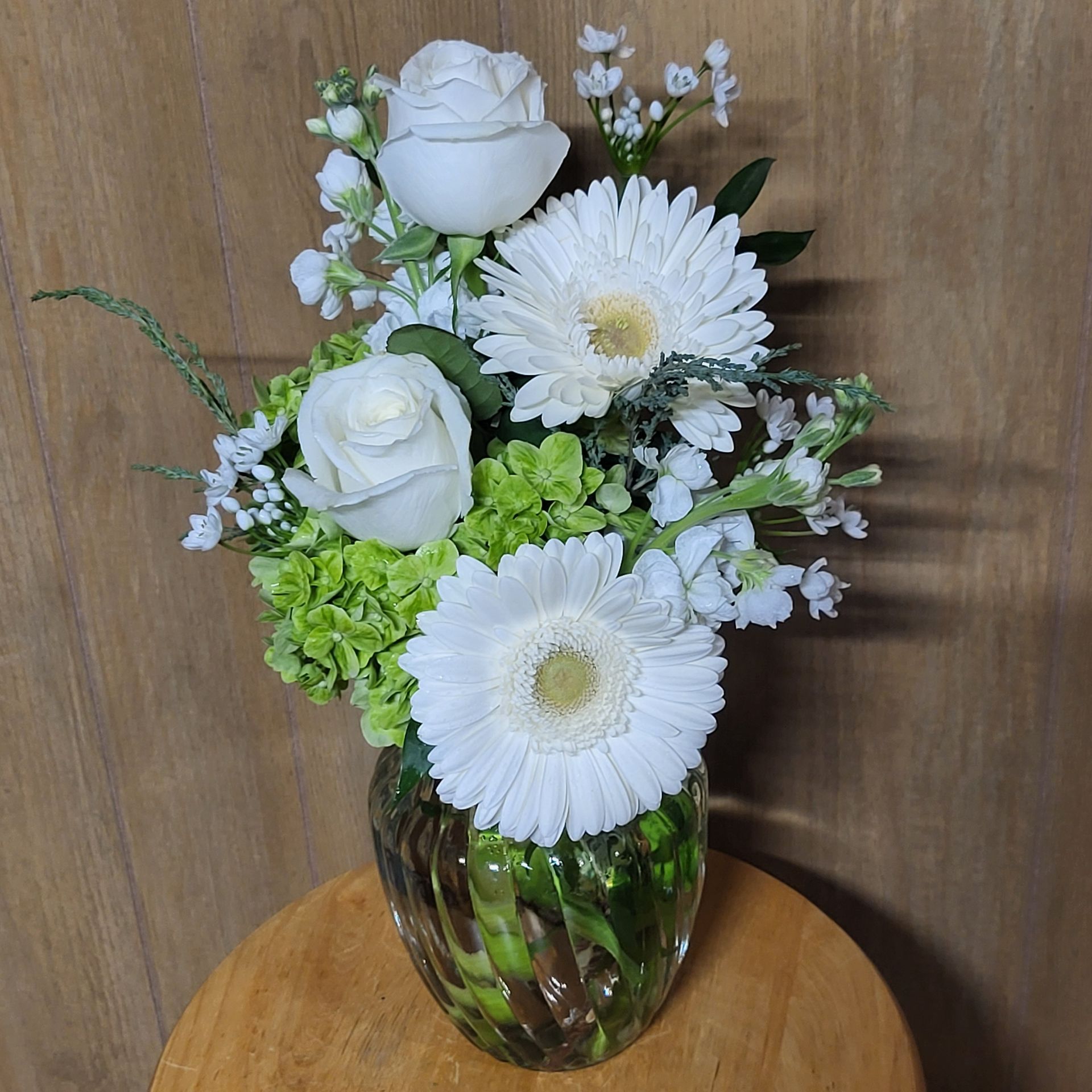 A vase filled with white flowers and greenery is sitting on a wooden table.