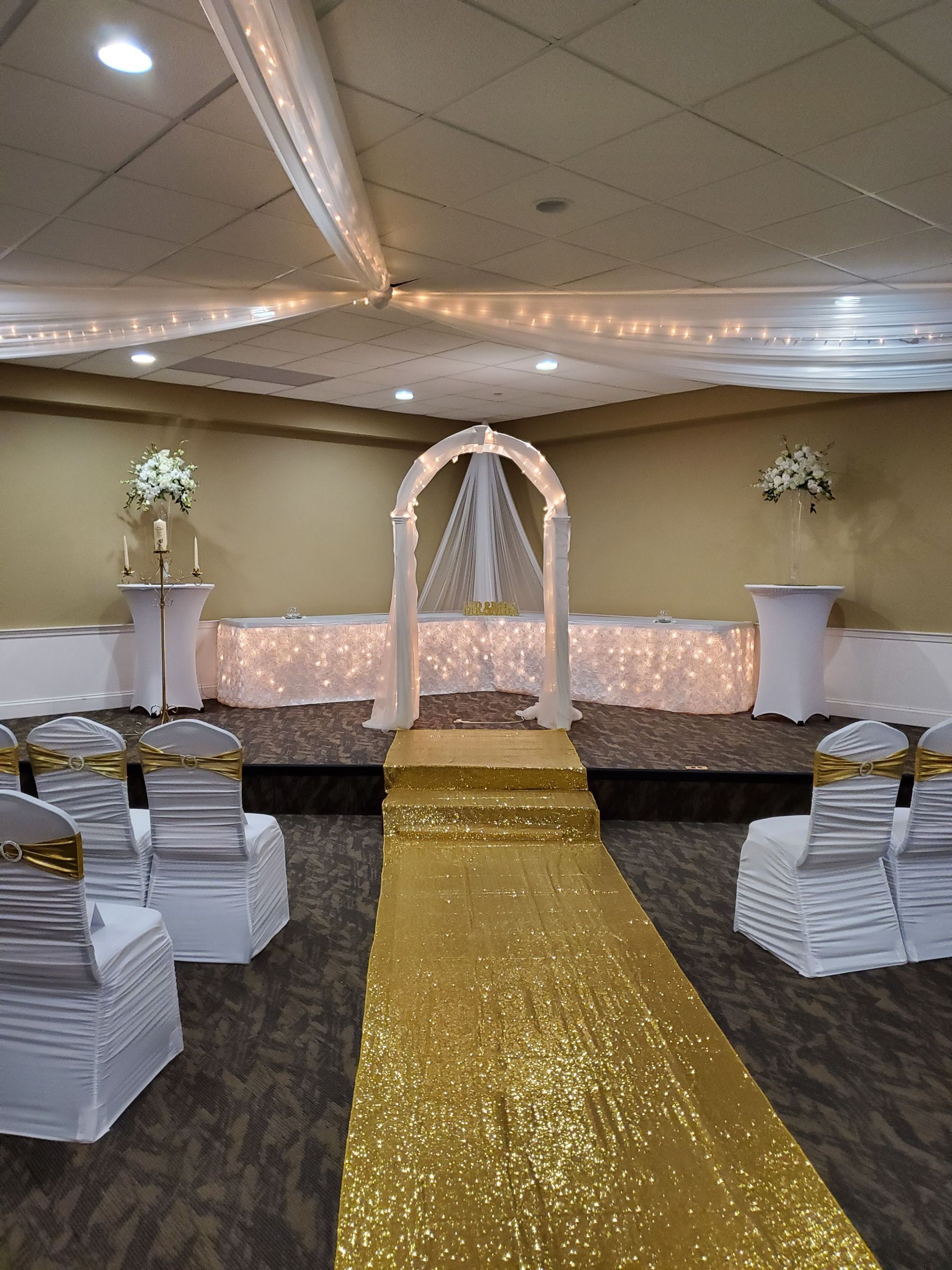 A wedding ceremony is taking place in a room with chairs and a gold aisle.