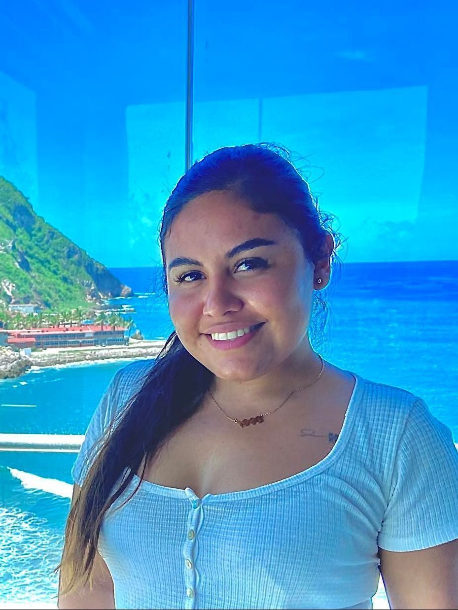 Woman smiles, ocean view through window. She wears a white top and a necklace.