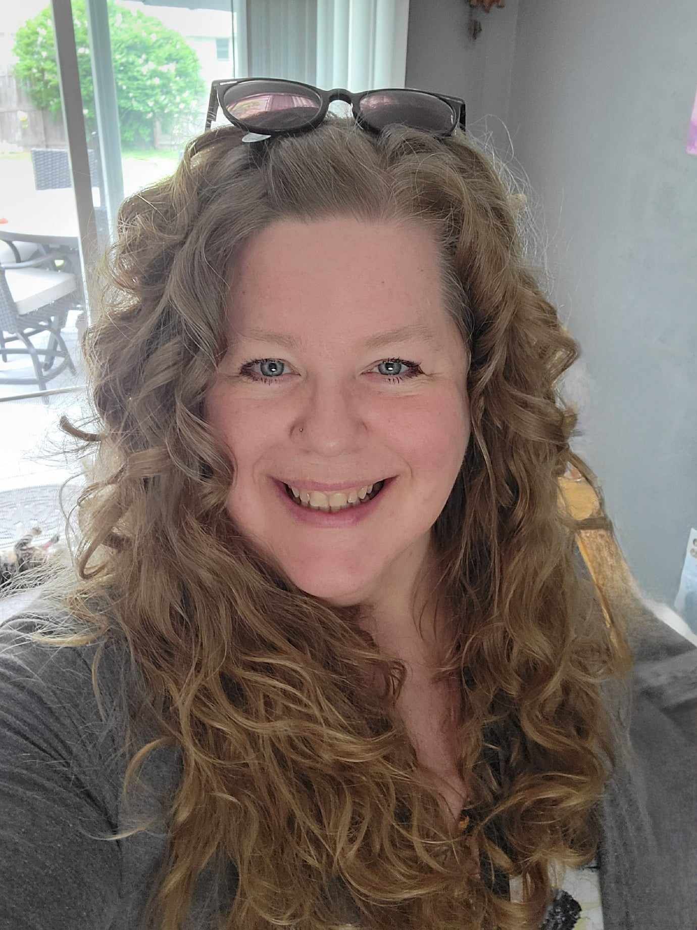 Woman with curly hair smiles, glasses on head, near a window.