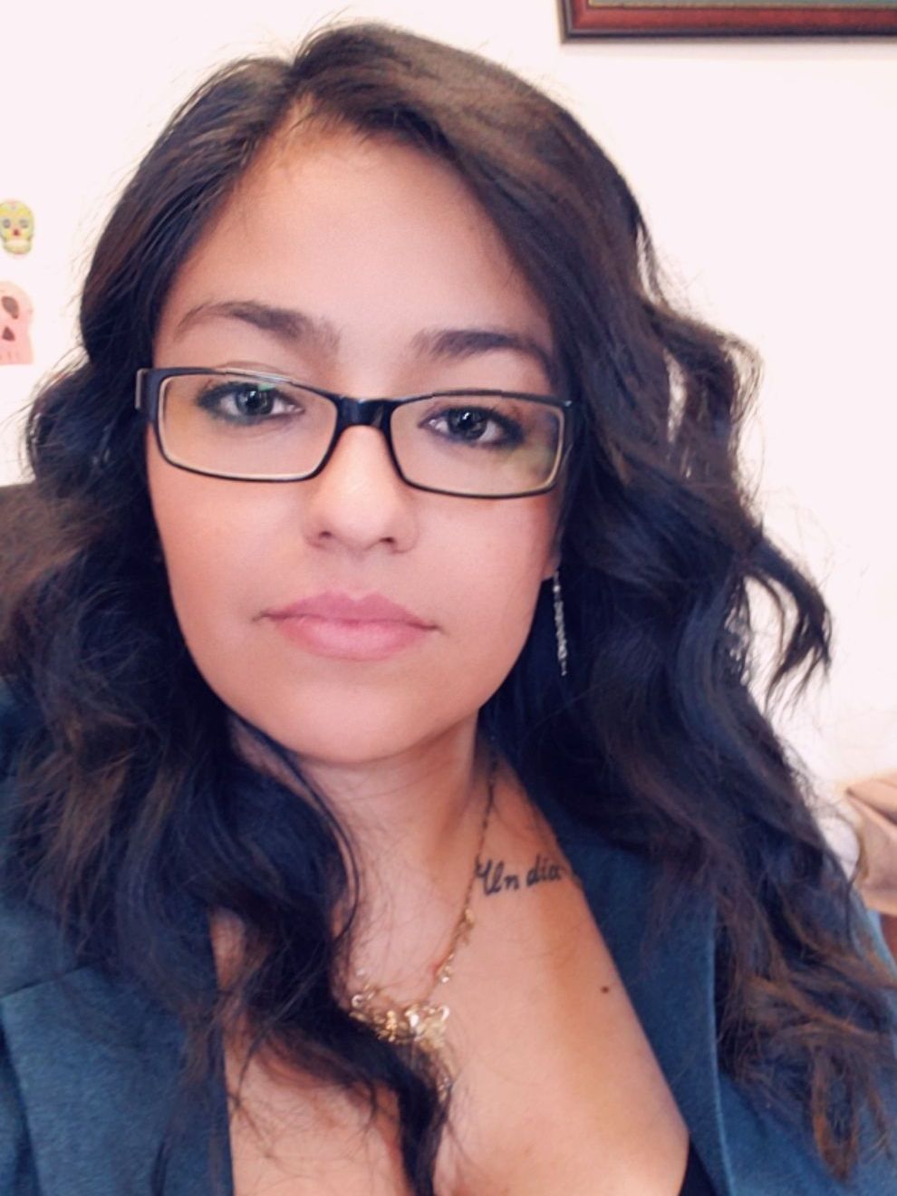 Woman with dark wavy hair and glasses, wearing a blazer. She has a pendant necklace and a small neck tattoo.