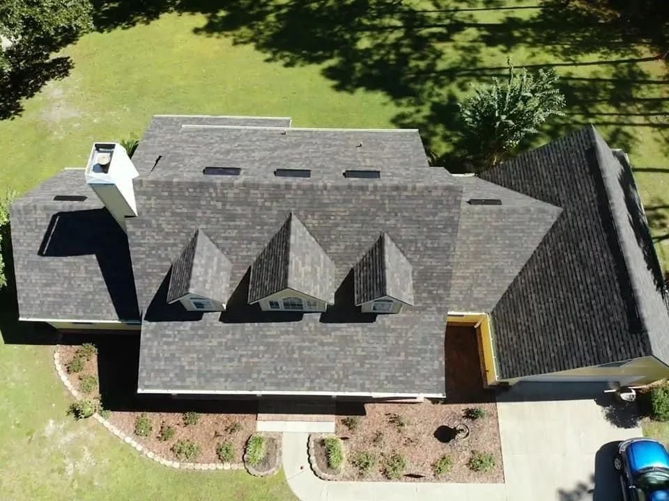 Aerial view of a residential house roof with three dormers, a chimney, and a landscaped yard, viewed from directly above.