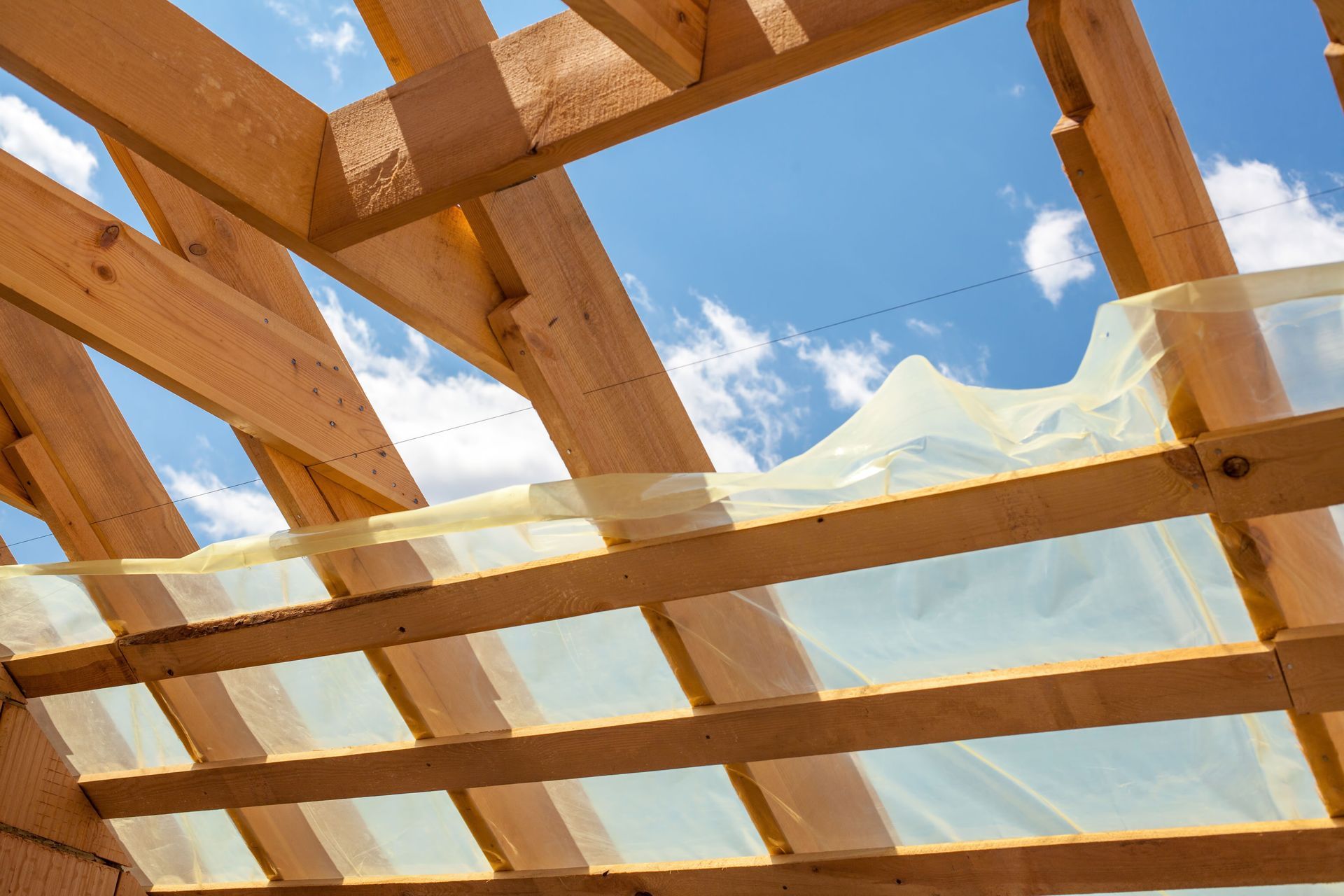 Wooden roof rafters of a building under construction, framed against a blue sky with thin, translucent plastic sheeting.