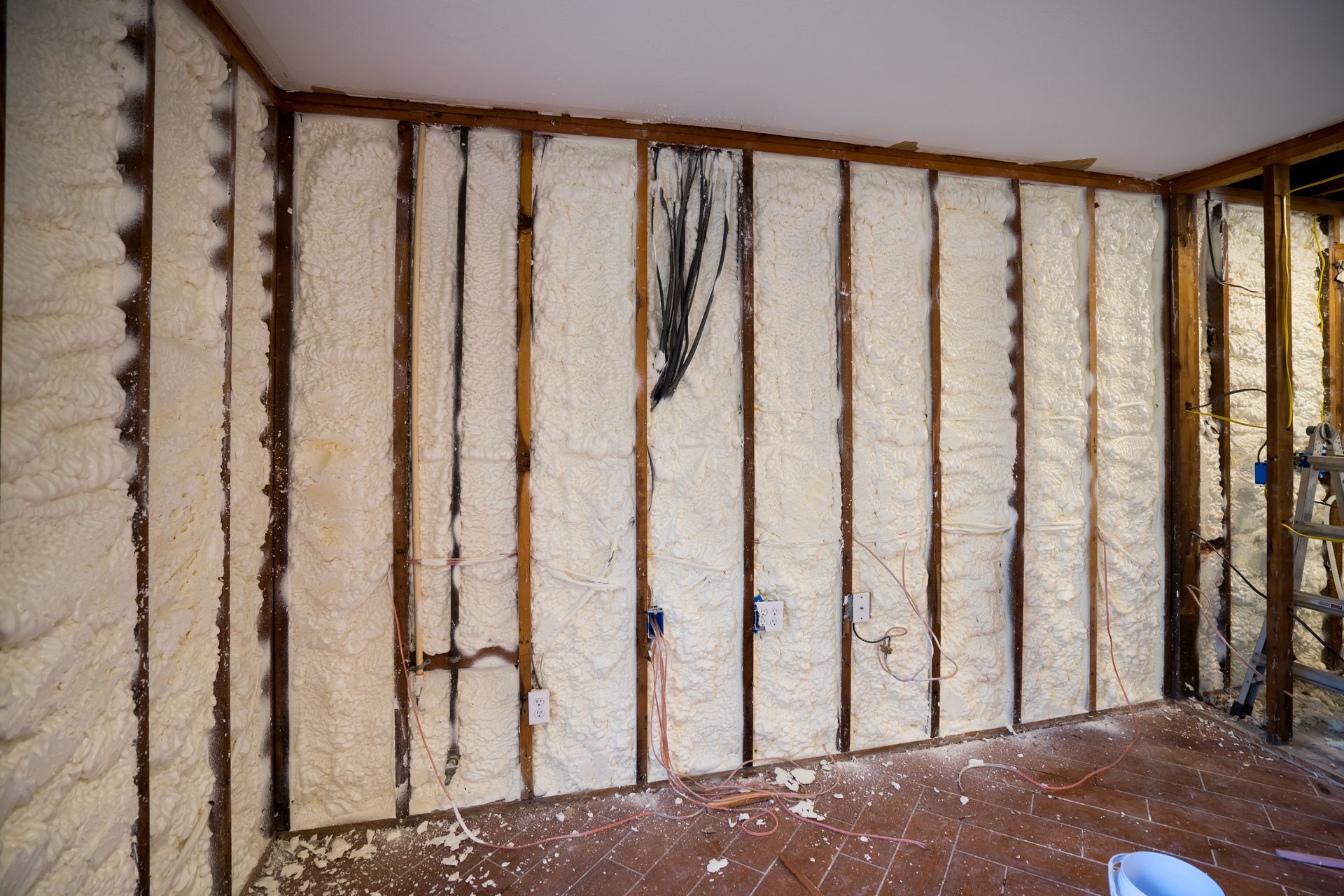 Interior view of a wall frame insulated with off-white spray foam, featuring exposed wiring and electrical boxes.
