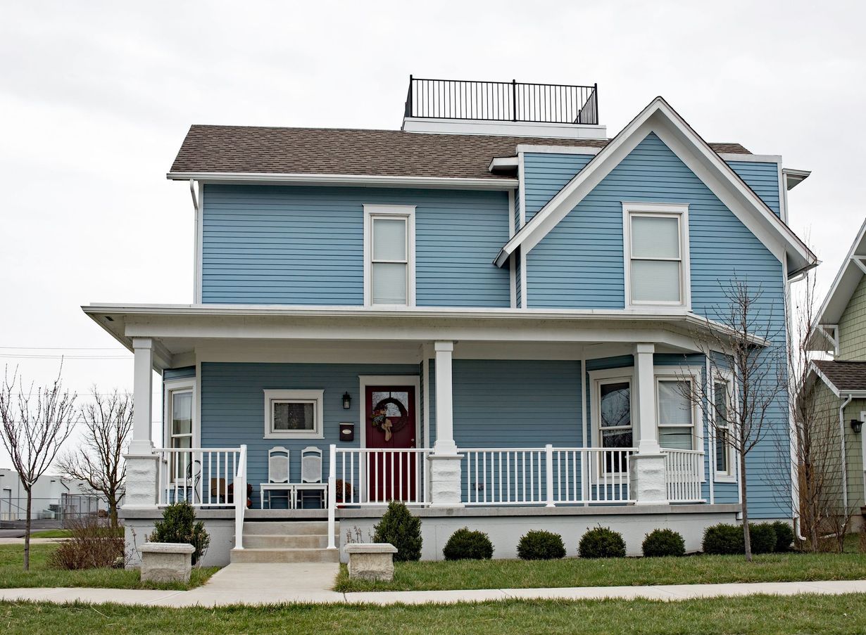 Blue two-story house with porch, white railings, and rooftop deck, set on green lawn.