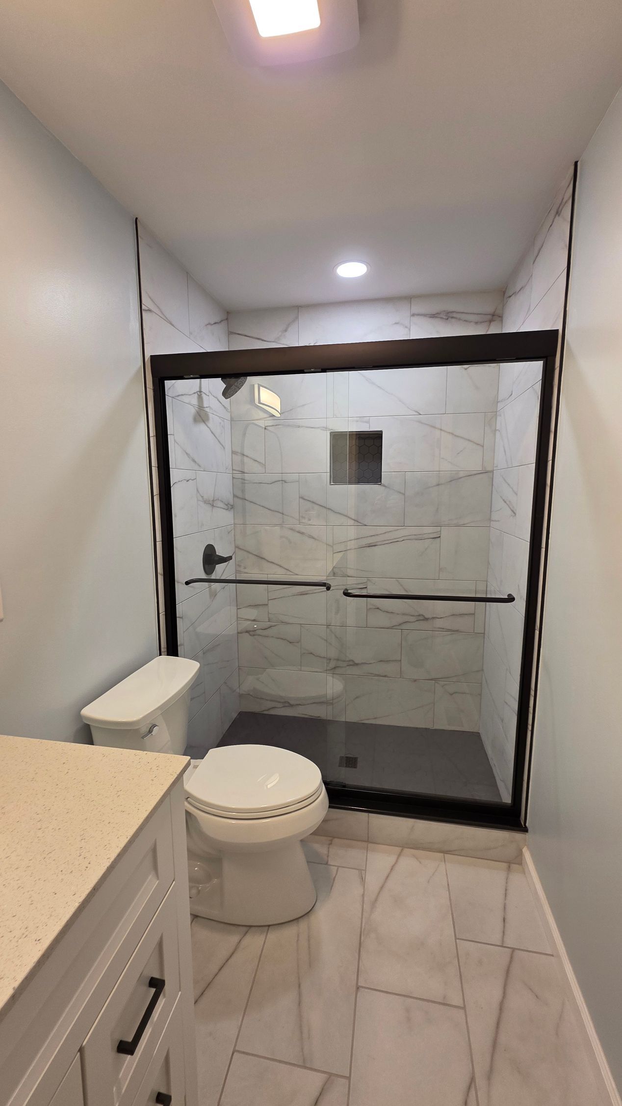 Bathroom with a shower, toilet, and vanity. White tile floors and walls. Dark-framed shower door.