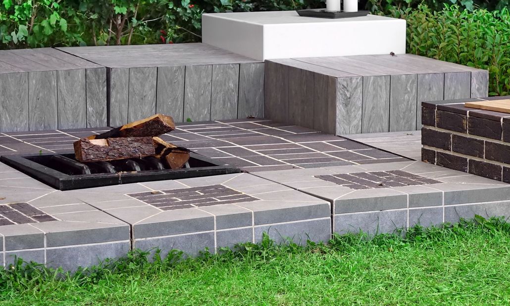 A patio fire pit area with grey stone pavers, a metal fire grate with logs, and a stepped retaining wall on a lawn.