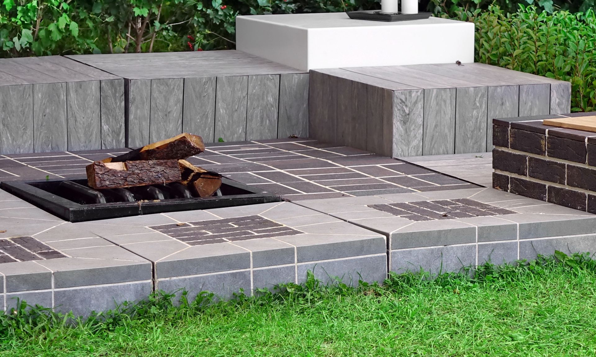 A patio fire pit area with grey stone pavers, a metal fire grate with logs, and a stepped retaining wall on a lawn.