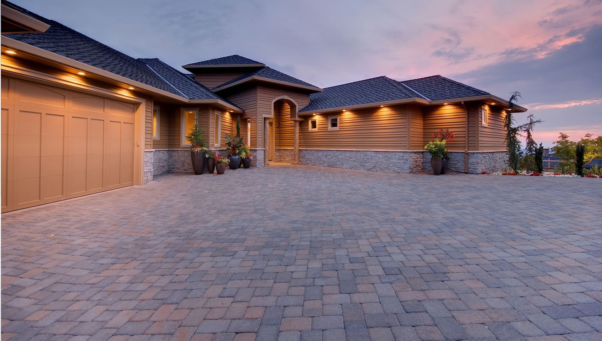 A modern home with a stone base, wooden siding, and a large paver driveway at dusk under a purple and pink sunset sky.