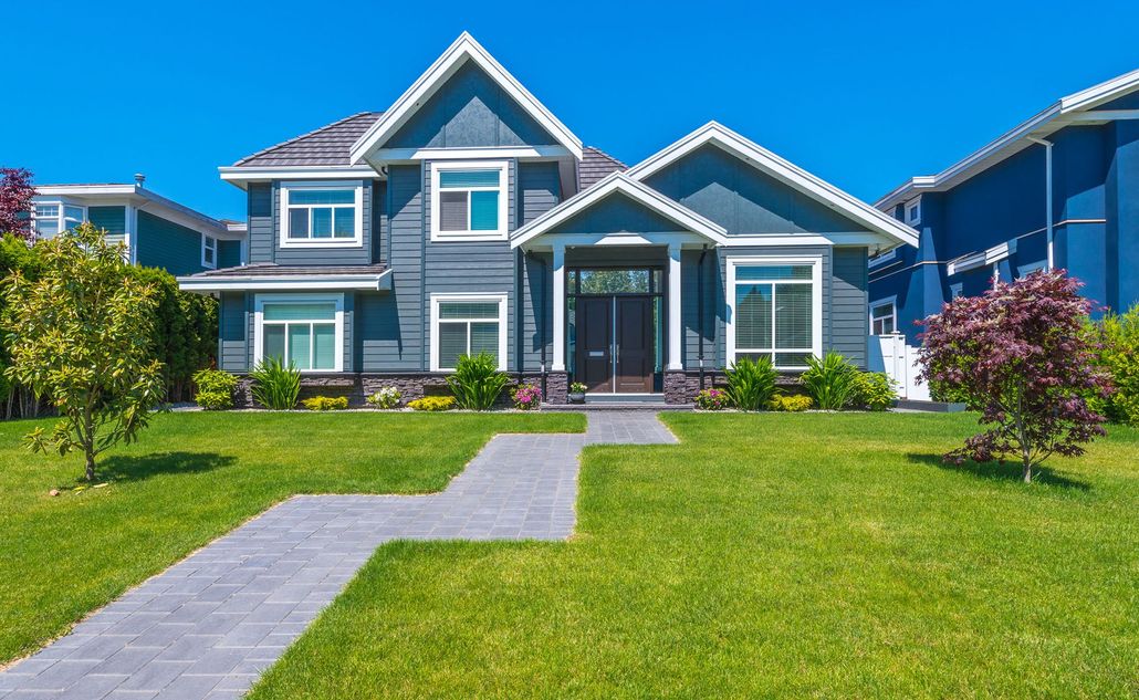A blue two-story suburban house with a paved walkway, green lawn, and clear blue sky.