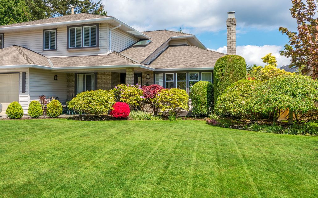 A two-story suburban house with beige siding, a shingled roof, and a well-manicured lawn with various shrubs.