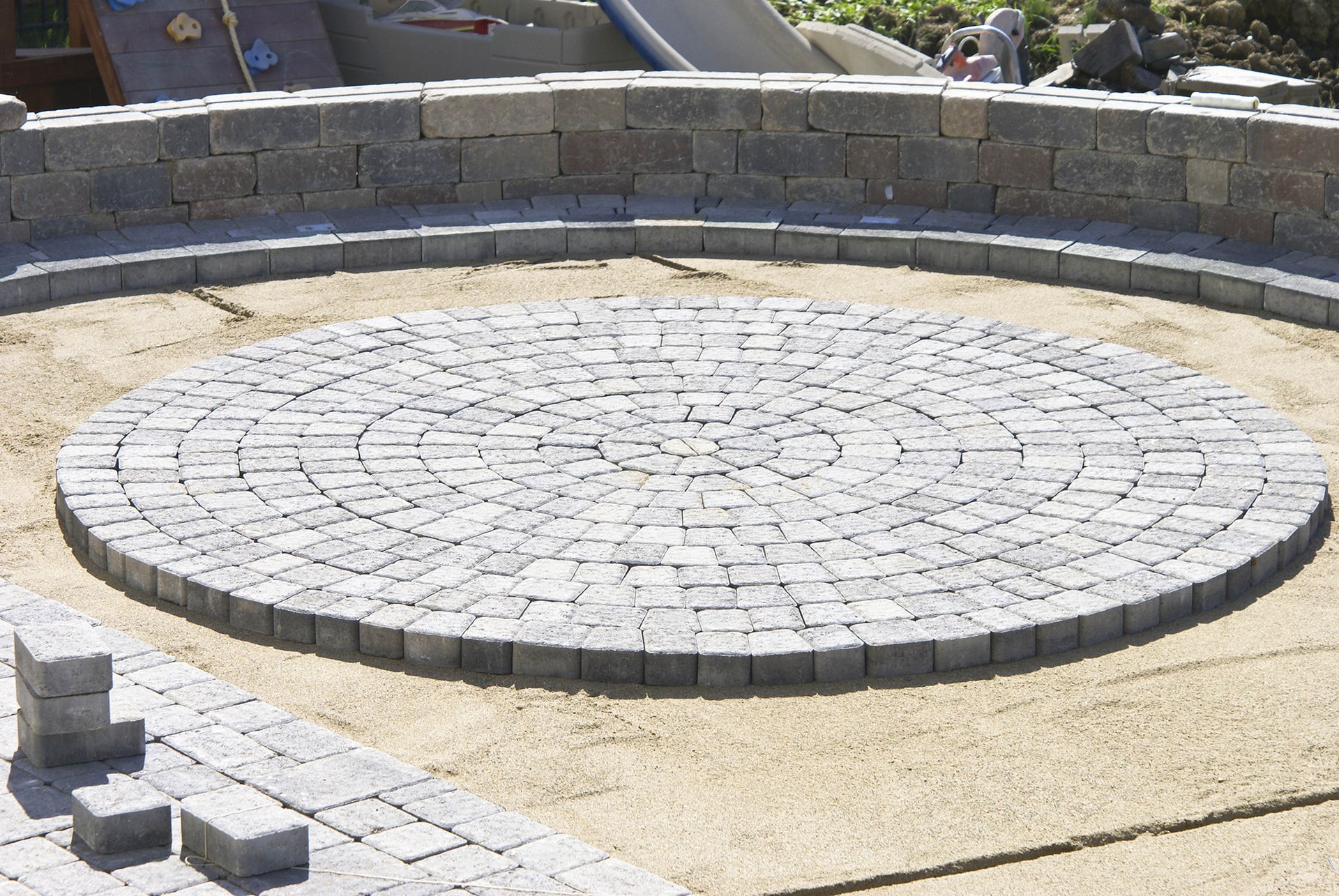 A circular stone patio being constructed, featuring gray pavers laid in concentric rings within a backyard setting.