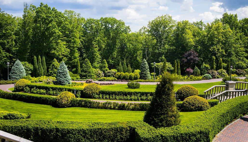 A manicured formal garden with tiered boxwood hedges, shrubs, and a path, backed by a lush forest under a cloudy sky.