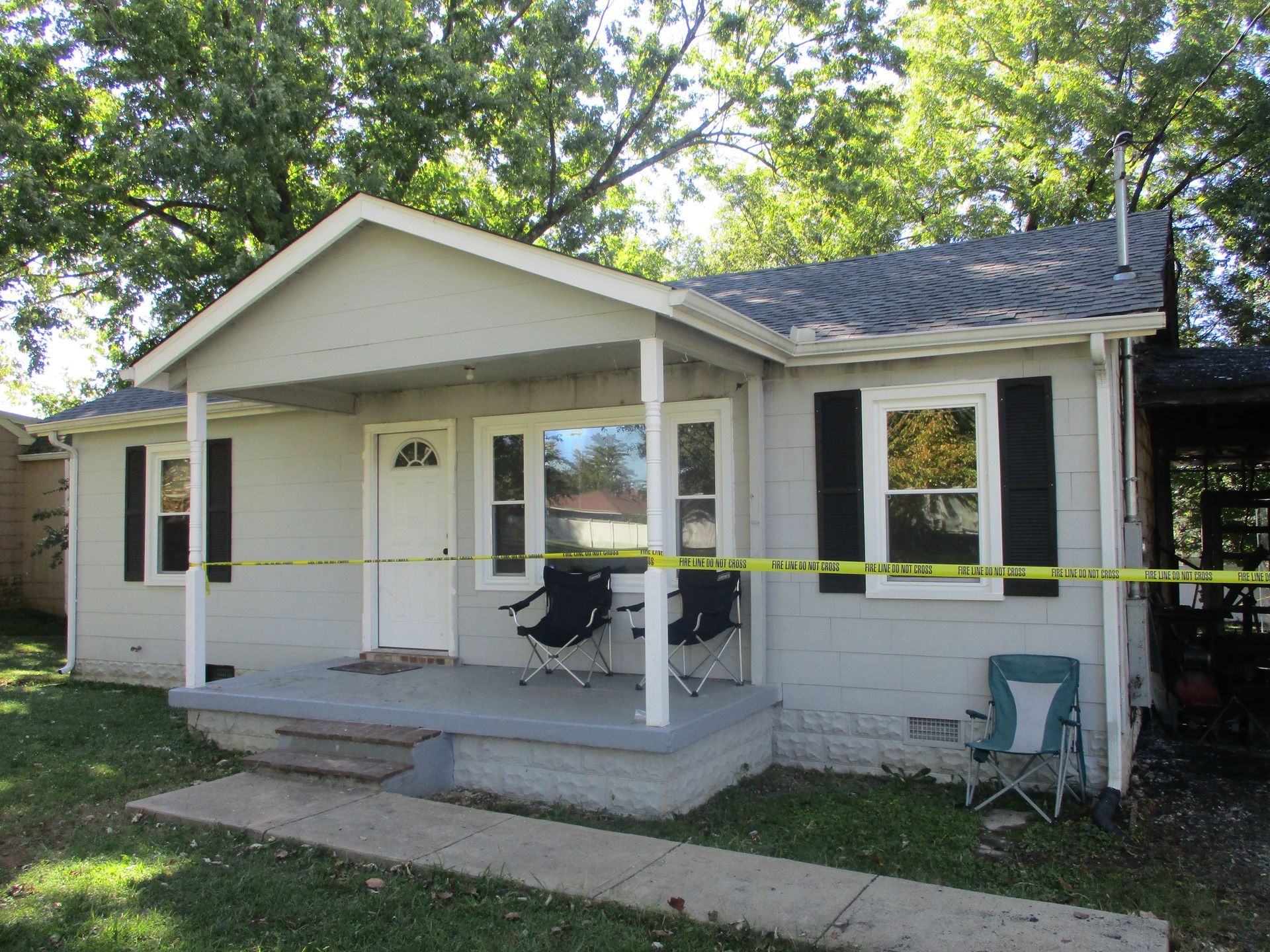 A small white house with a porch and black shutters is surrounded by yellow tape.