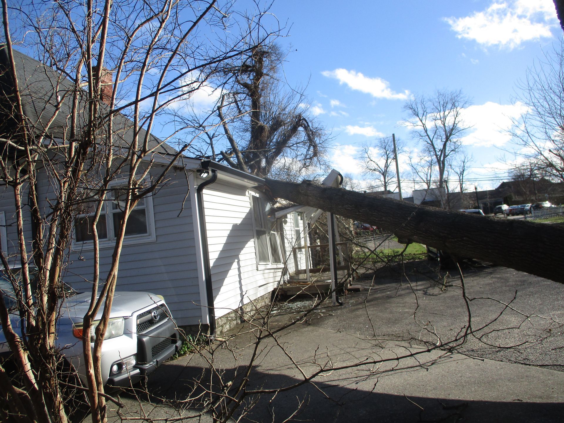 A car is parked in front of a house with a tree fallen on it.