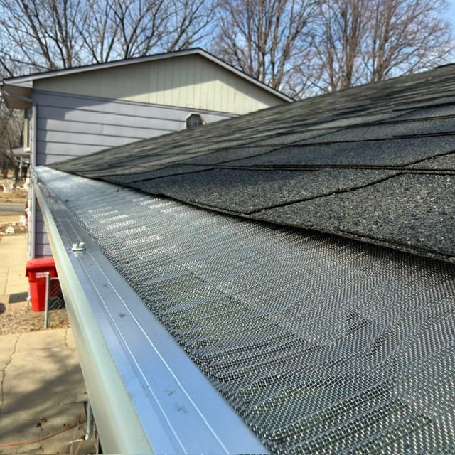 A silver metal gutter guard installed on a residential roofline with gray asphalt shingles.