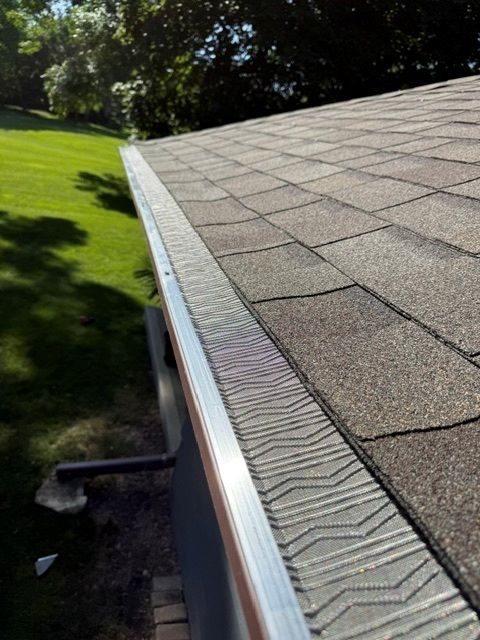 A close-up view of a shingled roof edge showing a metal gutter guard installed along the eave with a lawn in the background.