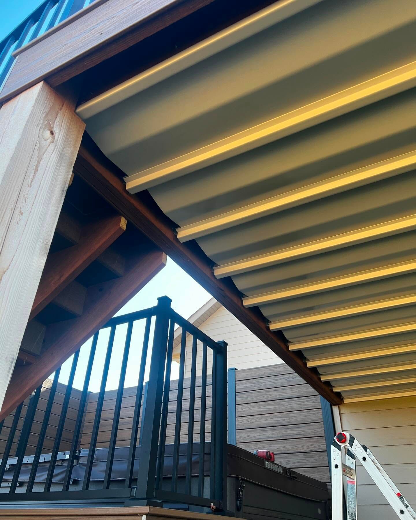 View of a deck's underside with a light-colored, corrugated ceiling, wooden beams, and a railing on stairs below.