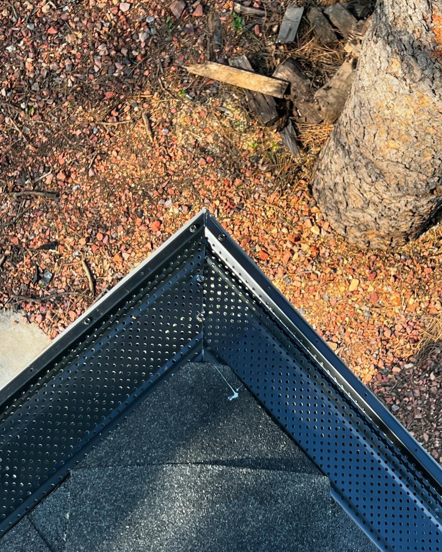 Black perforated gutter corner on a roof, next to a tree with brown mulch and debris.