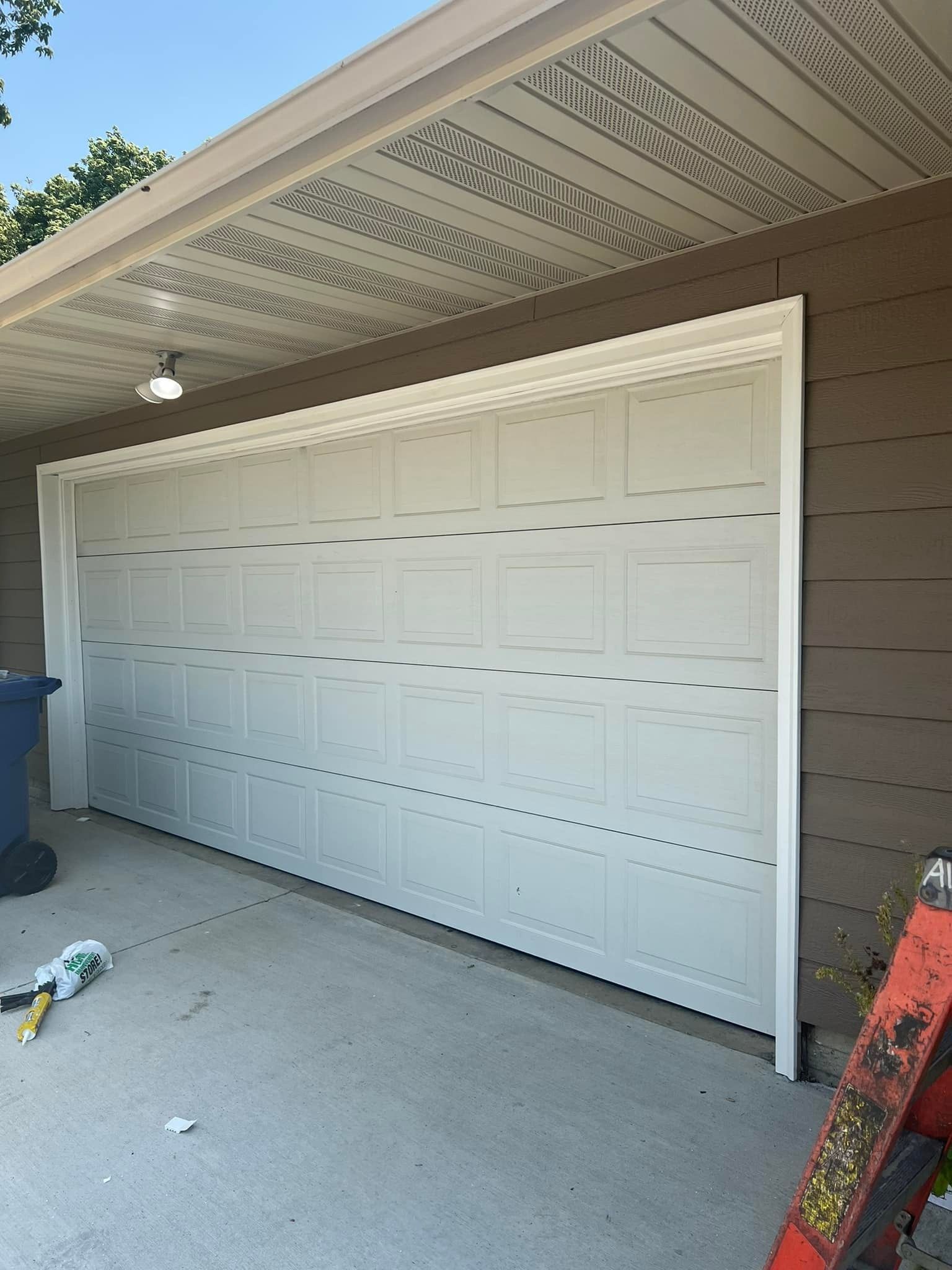 White garage door, partially open, under a brown roof. Ladder visible to the right, blue trash bin to the left.