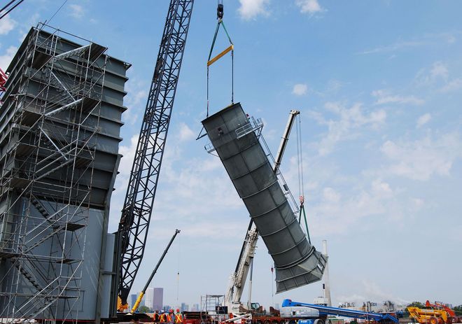 Construction cranes lifting a large curved concrete structure beside a high-rise under construction