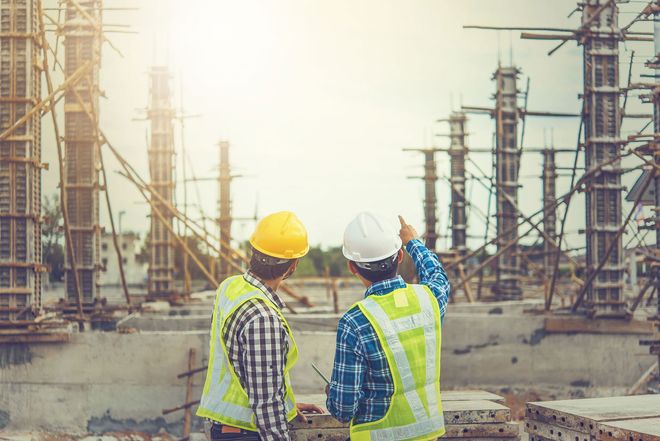 Two construction workers in hard hats inspect a large building site with scaffolding and concrete columns