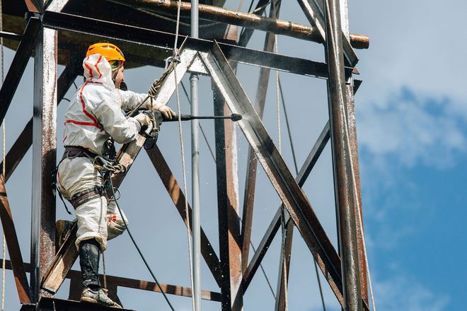 Worker in orange hard hat and safety gear climbing a metal tower against a blue sky
