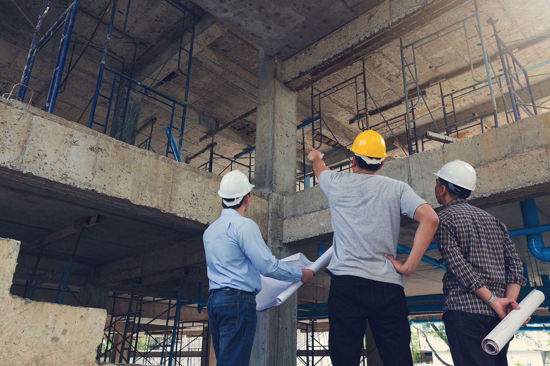 Three construction workers in hard hats inspect a concrete building structure at a job site.