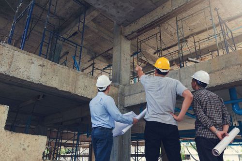 Three construction workers in hard hats inspect a building site with scaffolding and exposed concrete.