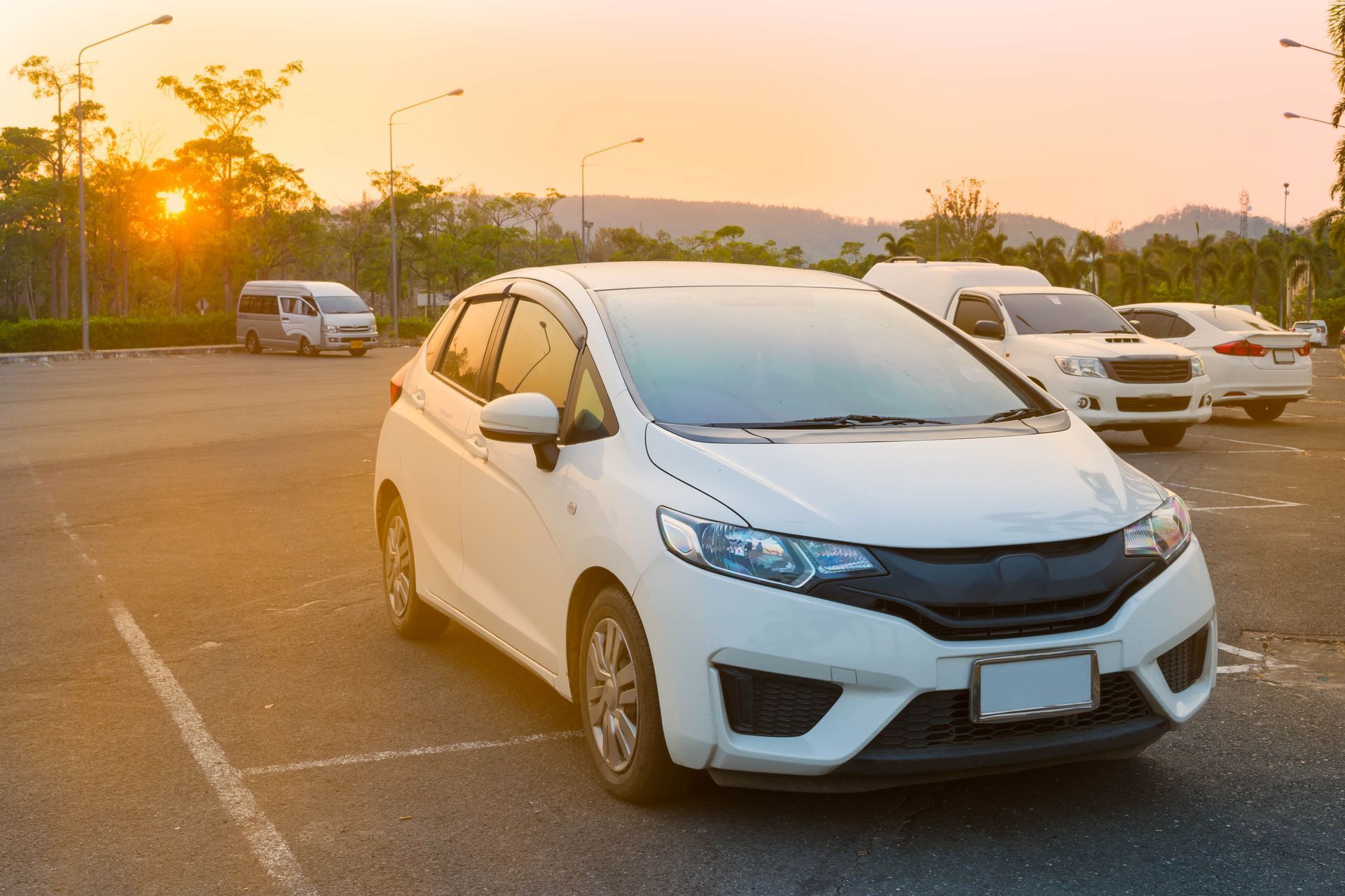 White car parked in a lot, other vehicles and trees in background with a sunset.