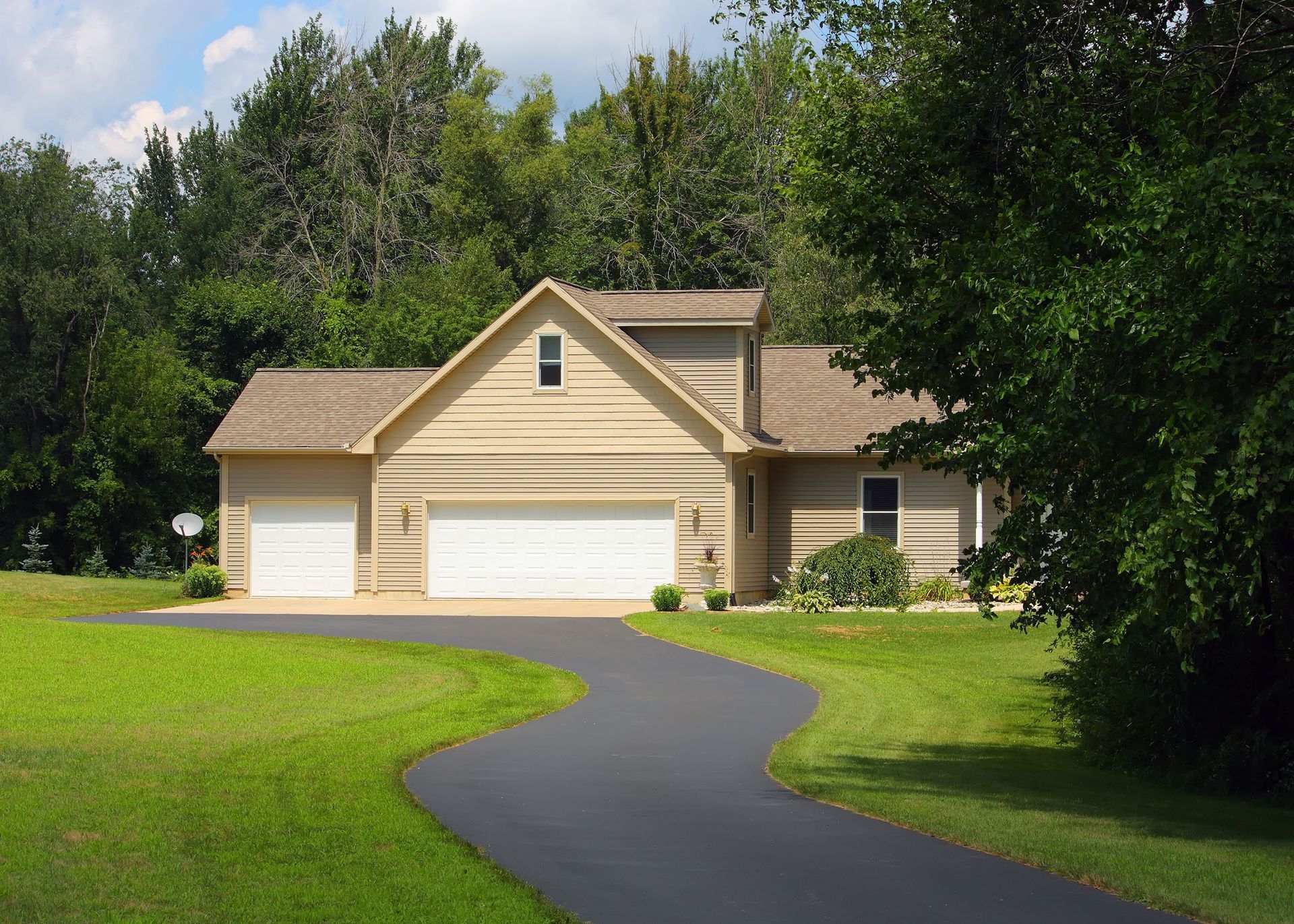 Tan house with two-car garage, winding driveway, and green lawn surrounded by trees.