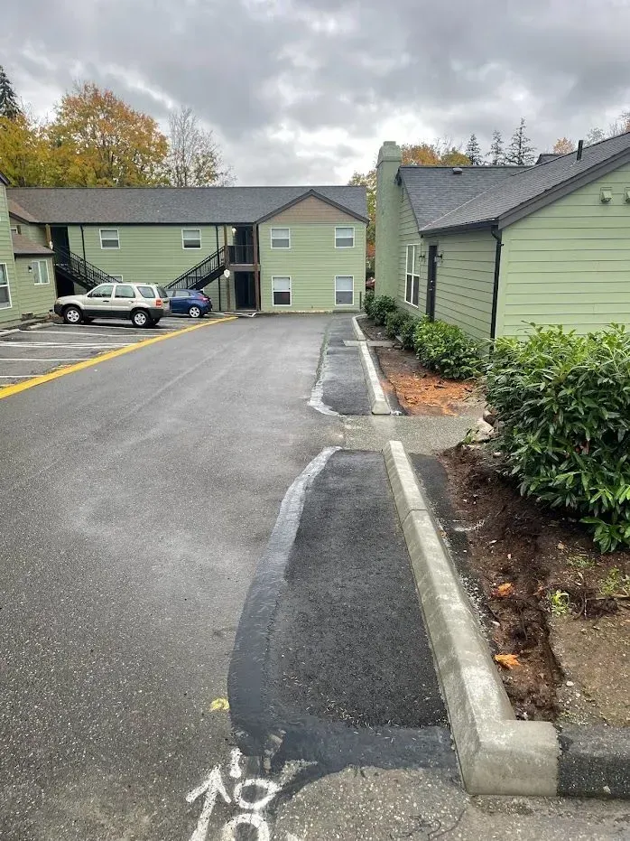 Apartment parking lot with freshly paved asphalt, curb, and green buildings under an overcast sky.