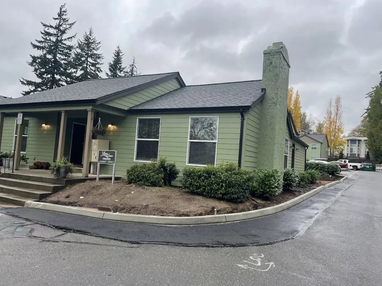 Green building with a dark roof and a tall chimney, next to a street with some trees.