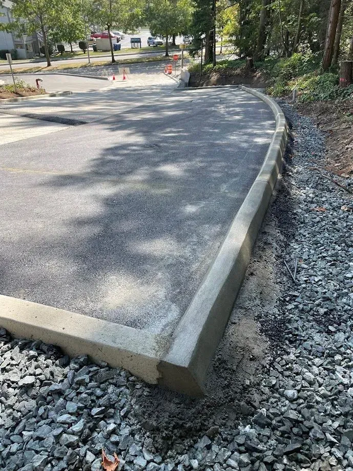 Asphalt parking area with concrete border, surrounded by gravel and trees.
