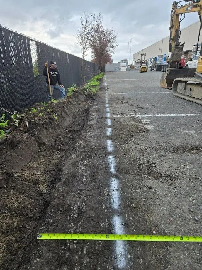 Man digging a trench next to a chain-link fence, a paved area with white lines, and an excavator.