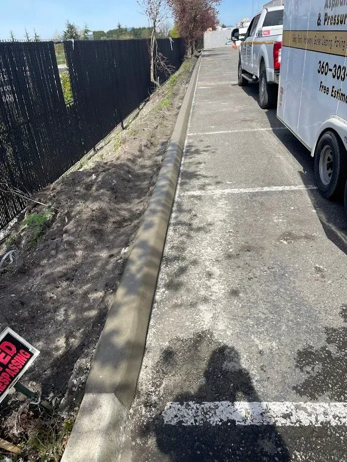 Concrete curb alongside a parking lot with a truck and trailer; a fence is on the left.