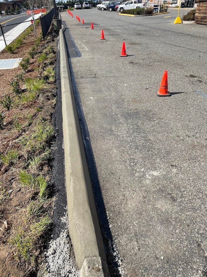 Concrete curb with orange cones, new landscaping on the left, and a parking lot with vehicles.