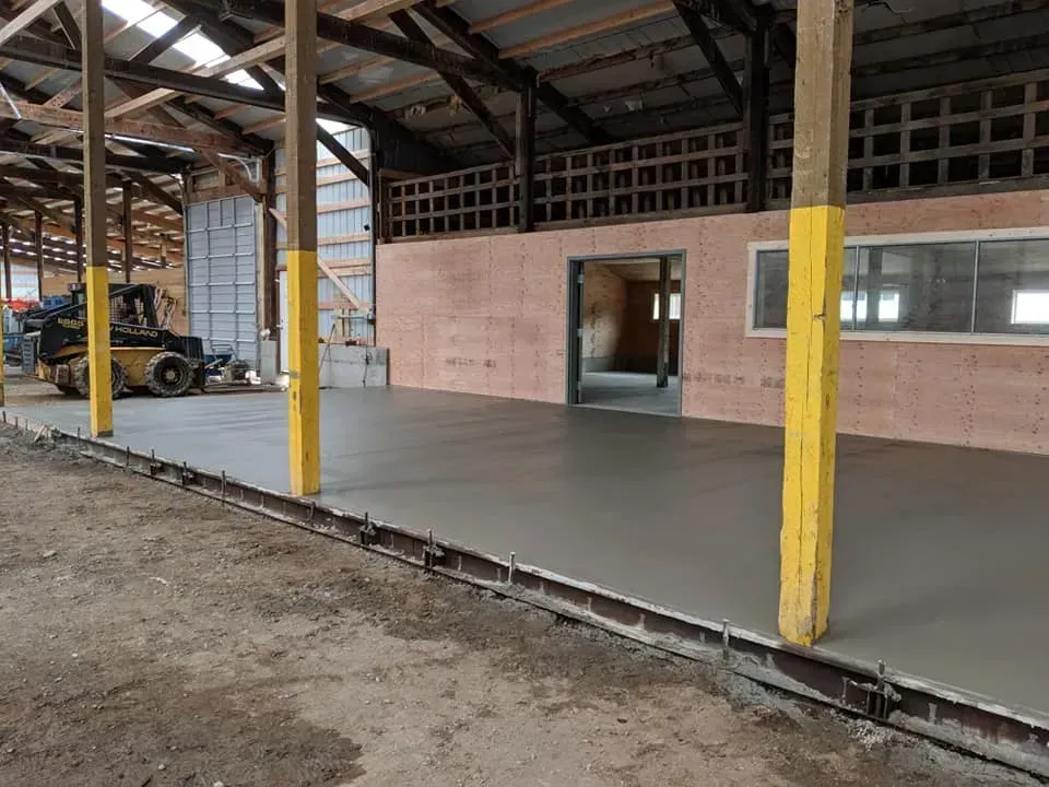 Interior view of a building with a newly poured concrete floor and yellow support beams. A small construction vehicle sits in the background.