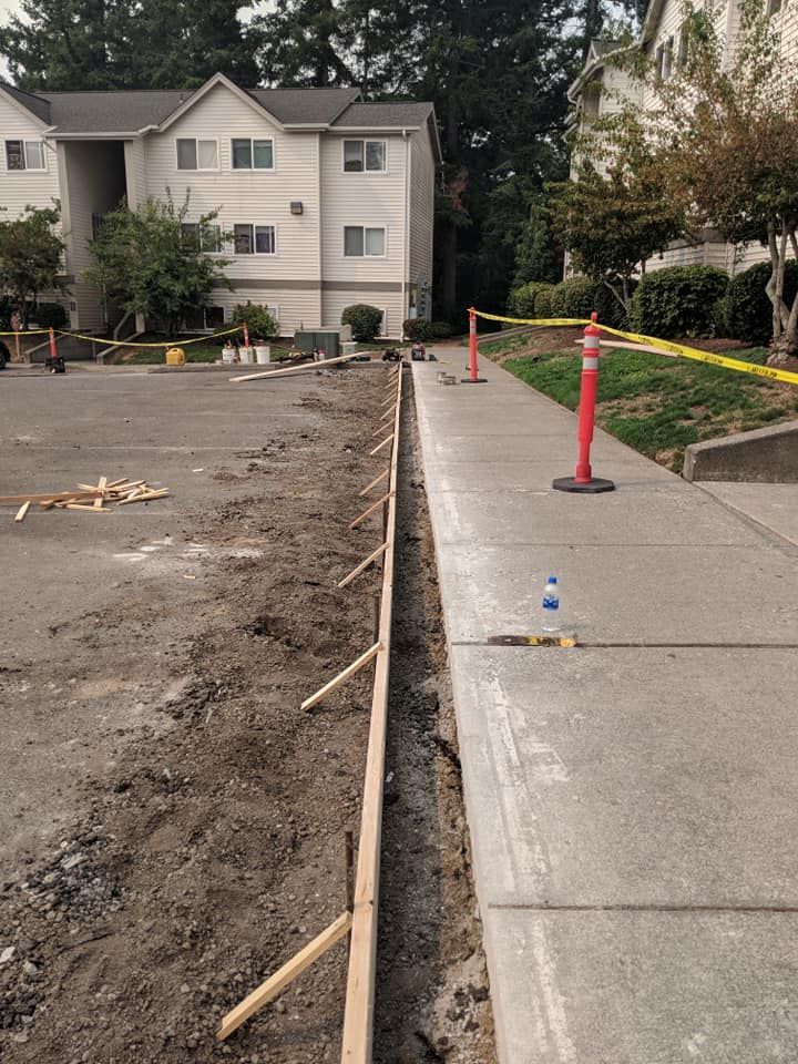 Construction site: concrete curb being built beside a sidewalk, with caution tape and apartment building in the background.