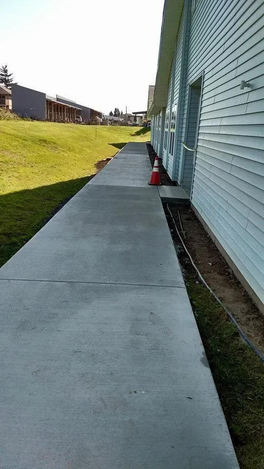Concrete sidewalk next to a building, with grass on the left. A traffic cone sits on the walkway.