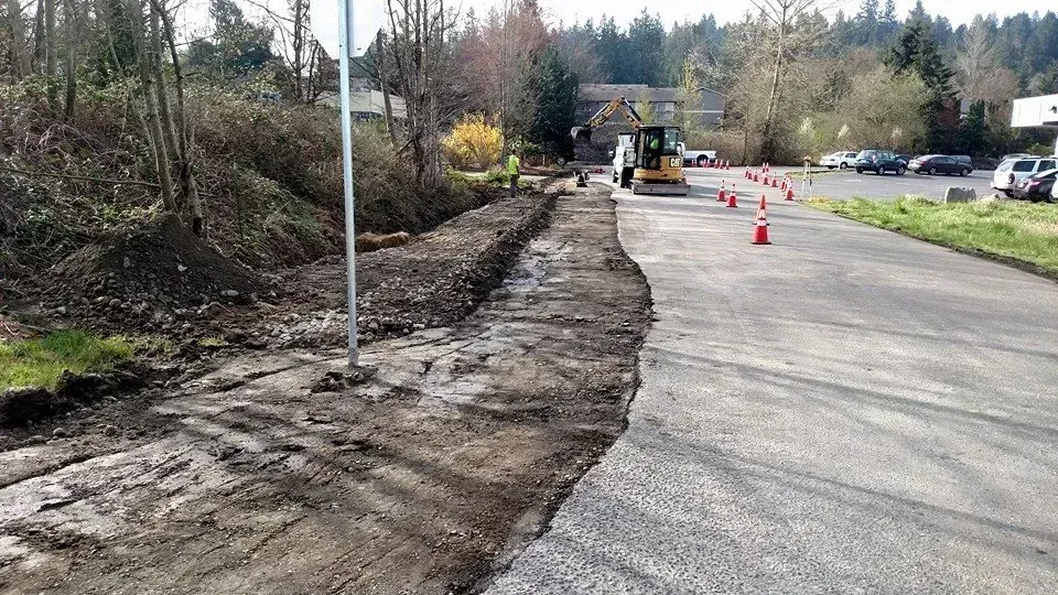 Road construction site with a ditch alongside a paved road, a mini excavator, and orange cones.