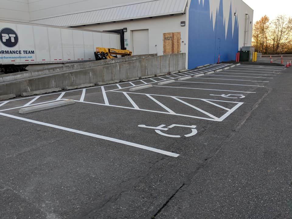 Parking lot with handicap spaces painted with white lines and symbols in front of a blue warehouse. A semi-truck is at a loading dock.