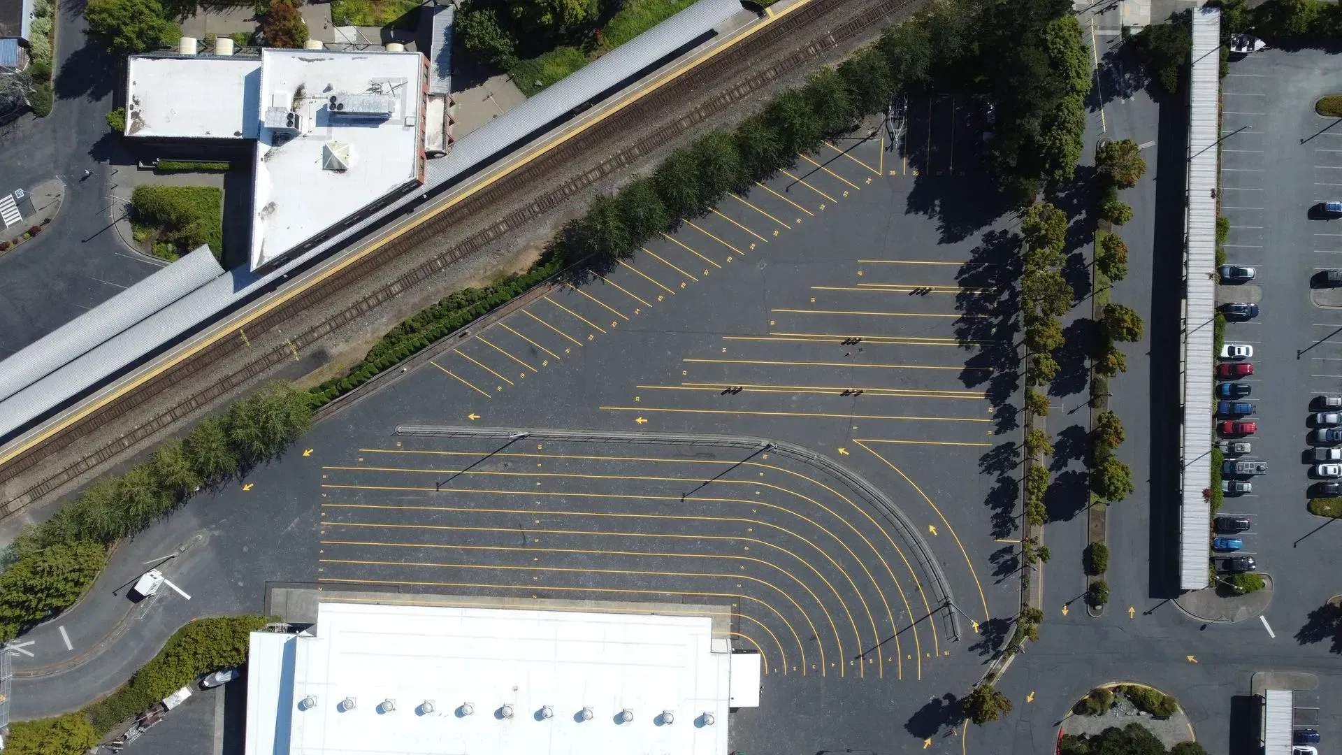 Aerial view of a parking lot next to a railway, building and trees.