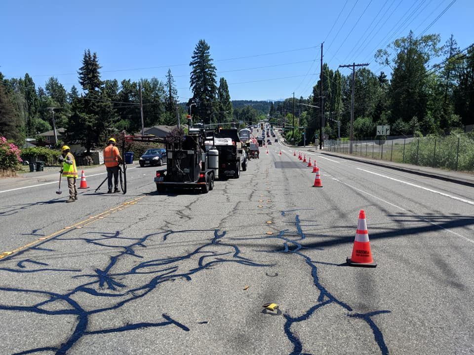 Road work on a cracked asphalt road with cones, trucks, and two workers in orange vests. Sunny day.