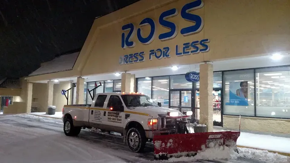 Snowplow clearing snow in front of a Ross Dress for Less store at night.