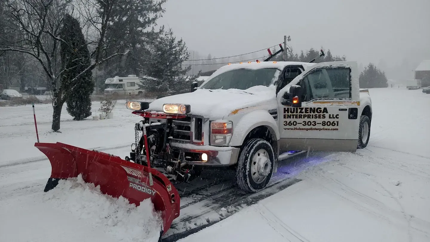 White snowplow truck clearing a snow-covered road in a residential area. The plow is red, and the sky is overcast.