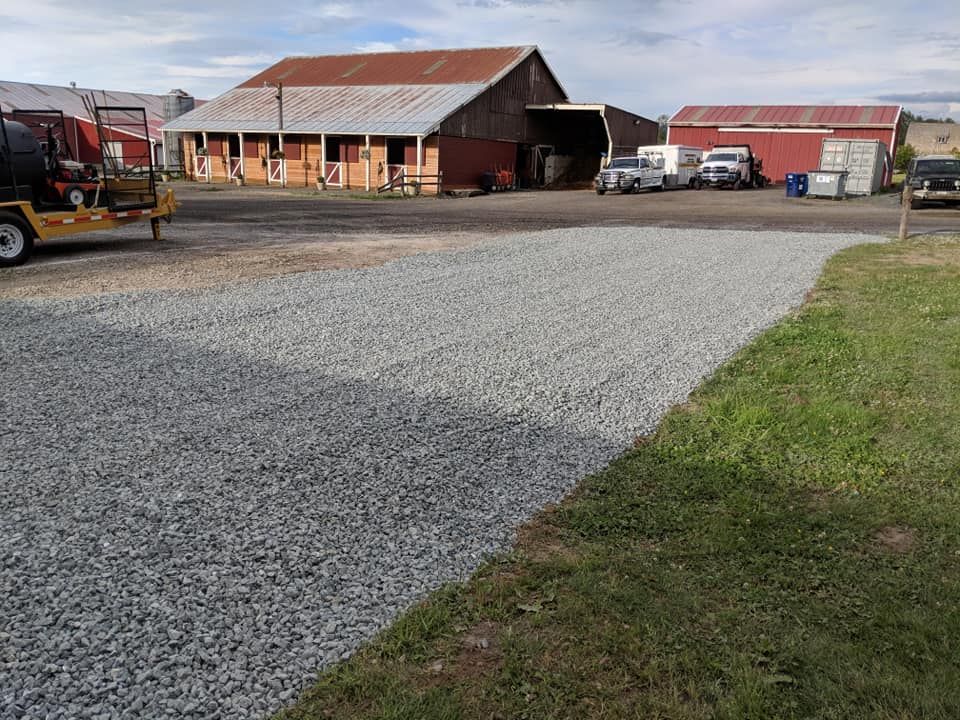 Gravel driveway next to a grassy area leading towards red barns and parked vehicles.