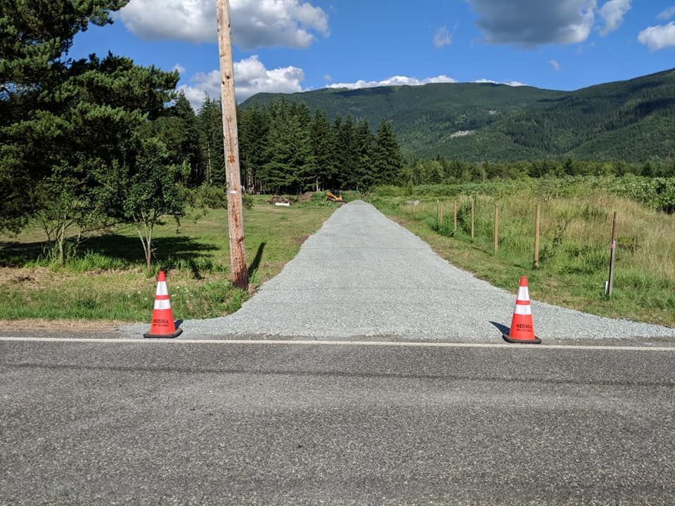 Gravel driveway from road, flanked by cones, leading to a field with mountains in the background.