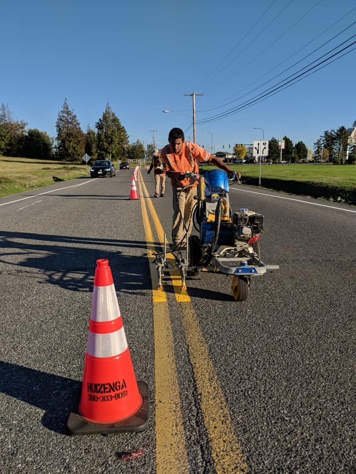 Man painting yellow double lines on an asphalt road with a line-marking machine, cones set out.